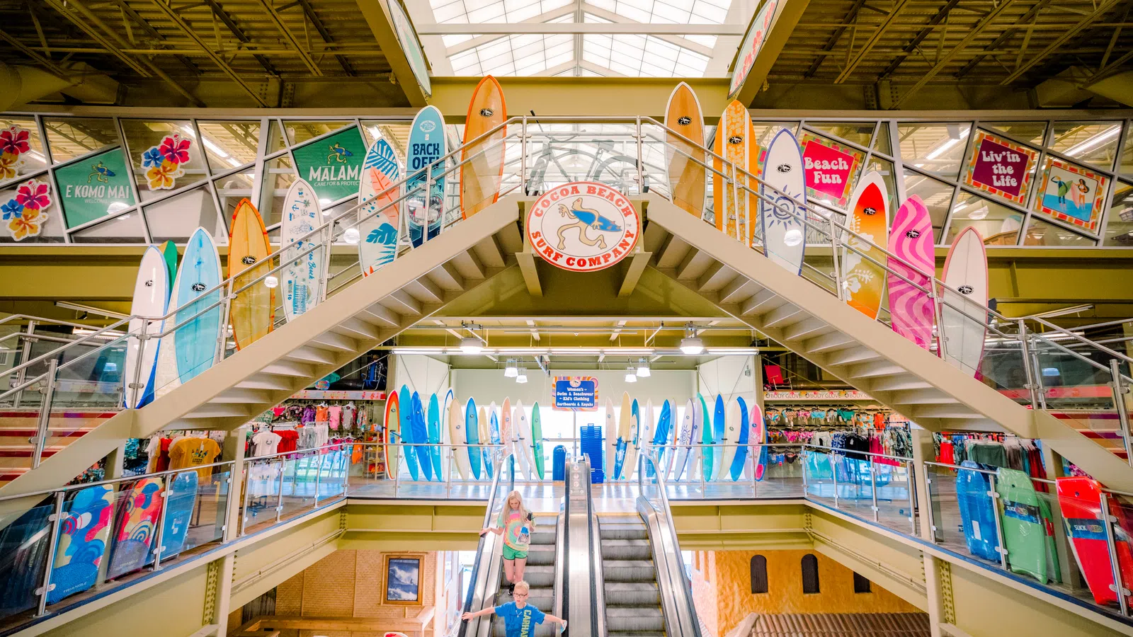 Interior of a surf shop with colorful surfboards displayed around a staircase and two people on an escalator.