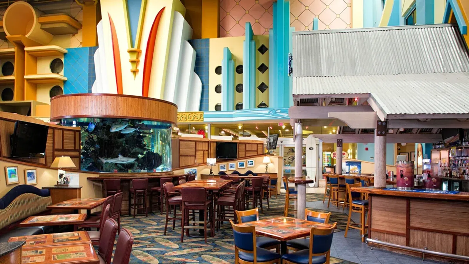 Colorful, retro-style restaurant interior with wooden tables, bar area, and large cylindrical fish tank centerpiece