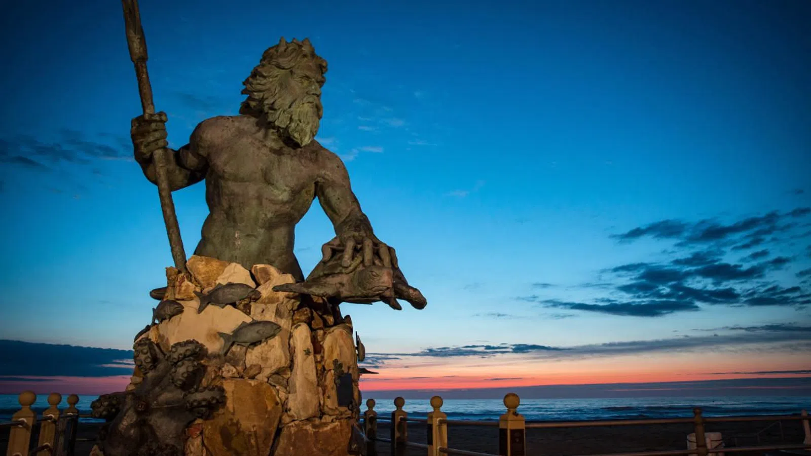 Statue of Poseidon holding a trident and sea creatures at sunset by the oceanfront.