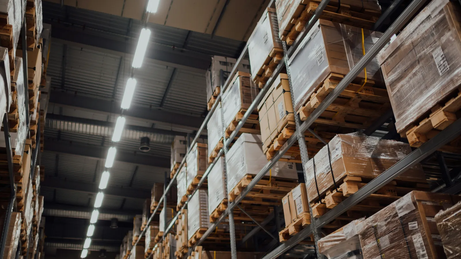 High warehouse shelves filled with pallets of wrapped boxes under bright industrial lighting in a large storage facility
