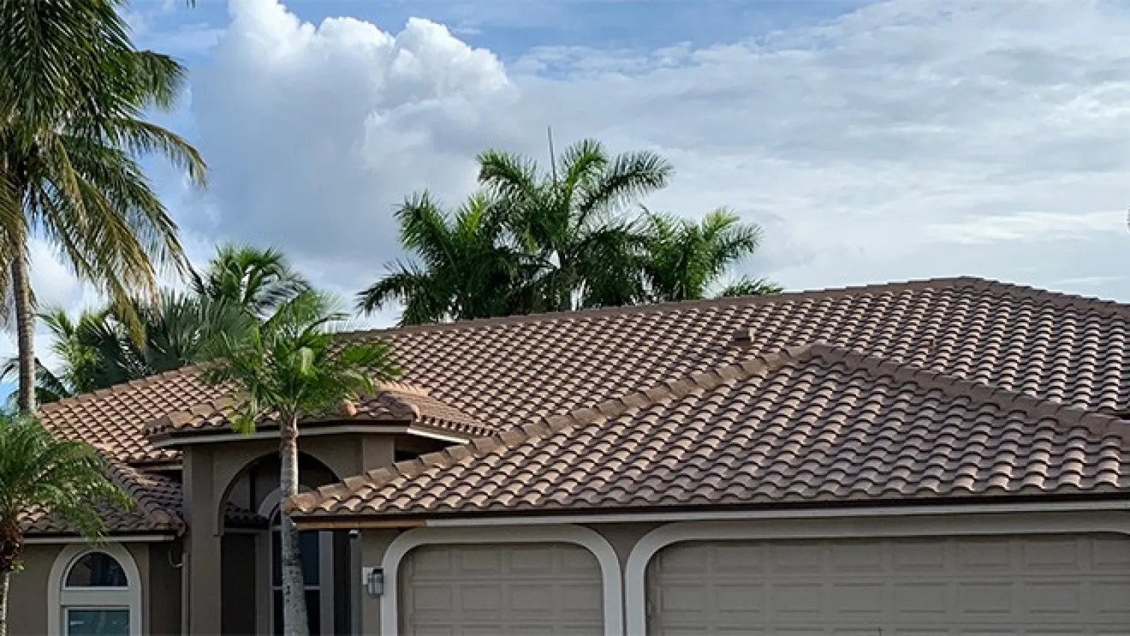Modern house roof with brown tiles and palm trees under a cloudy sky in a tropical setting