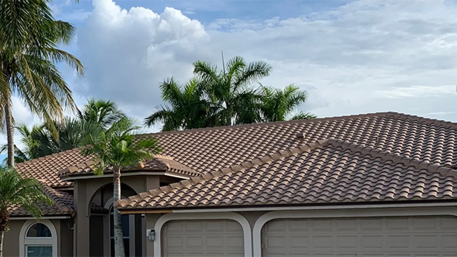Brown tiled roof on modern house with palm trees under a cloudy blue sky.