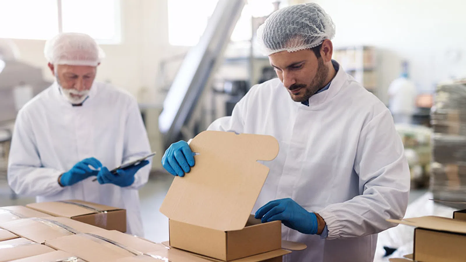 Two workers in white protective clothing packing products into cardboard boxes in a factory setting.