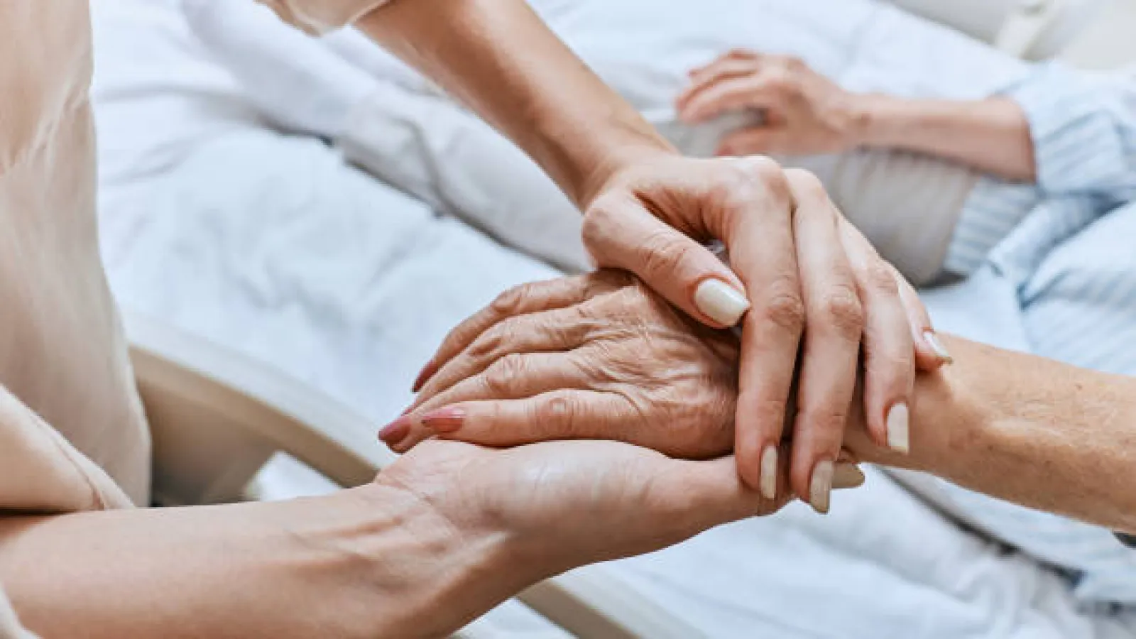 Younger hands gently holding elderly hands over a hospital bed, symbolizing care and support.