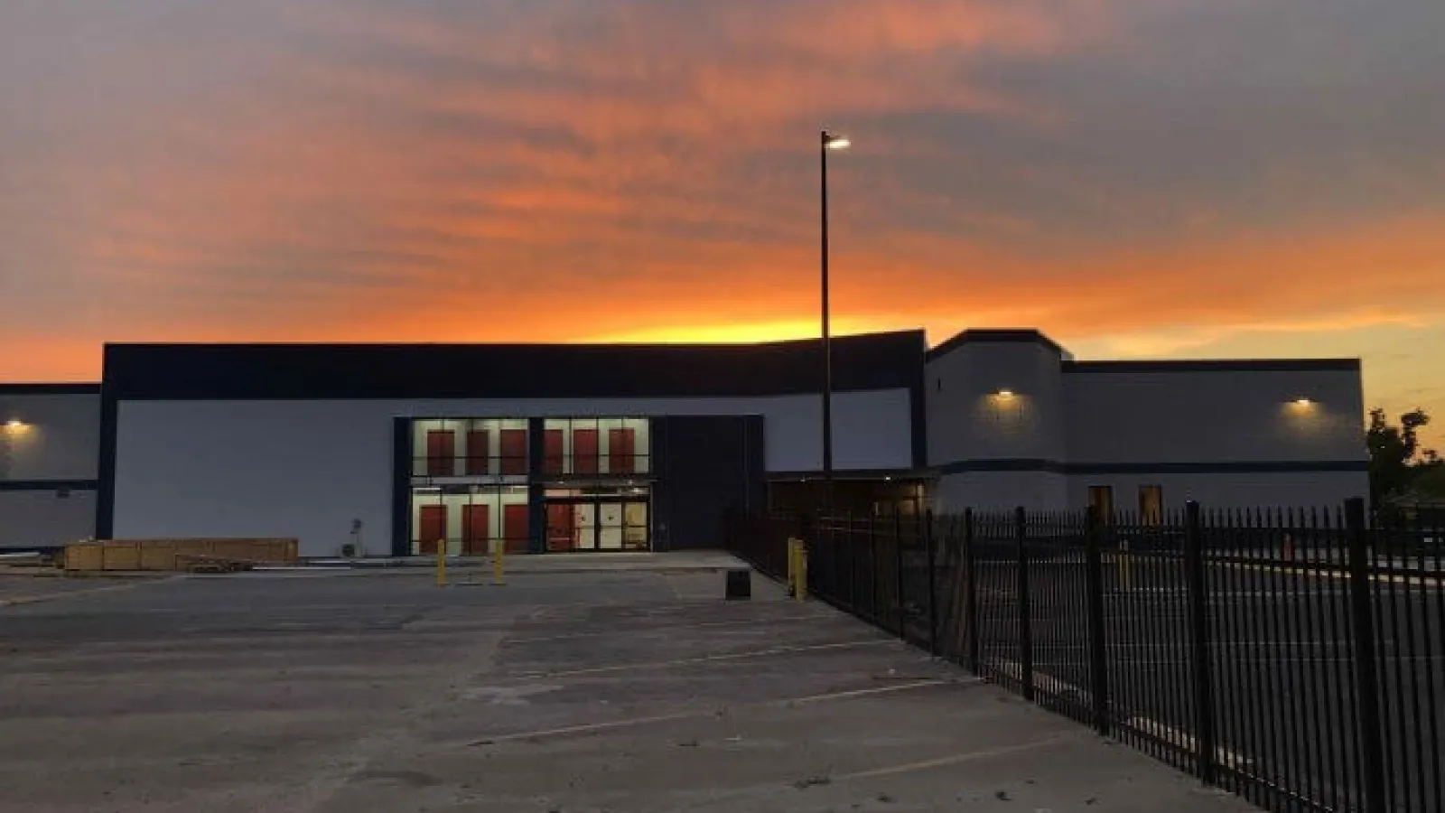 Modern industrial building with a parking lot and fence at sunset with vibrant orange sky.