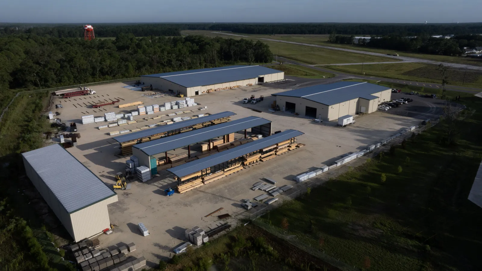 Aerial view of a lumber yard with storage buildings, wood stacks, and open loading areas surrounded by greenery.