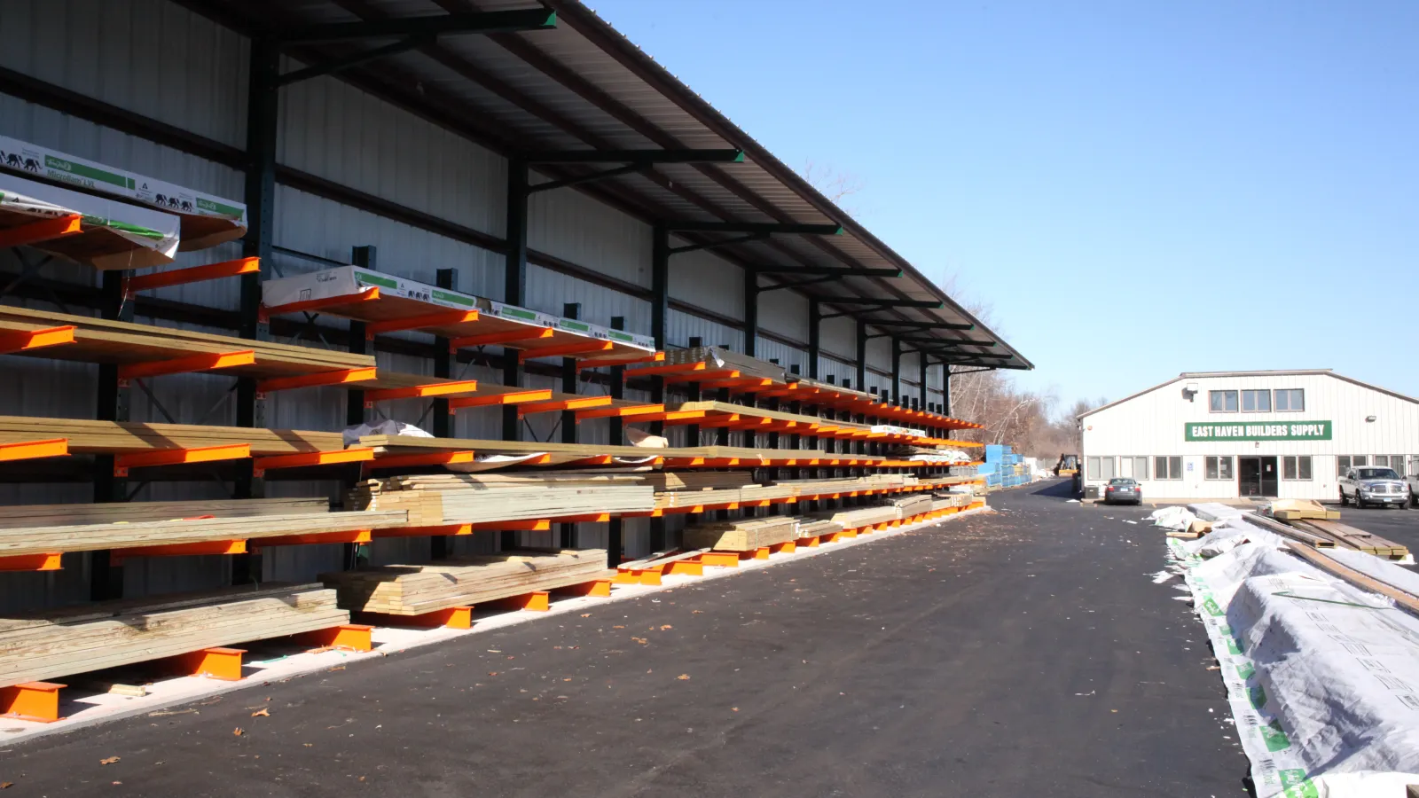 Outdoor lumber yard with stacked wood planks under a metal roof and a building in the background under clear sky