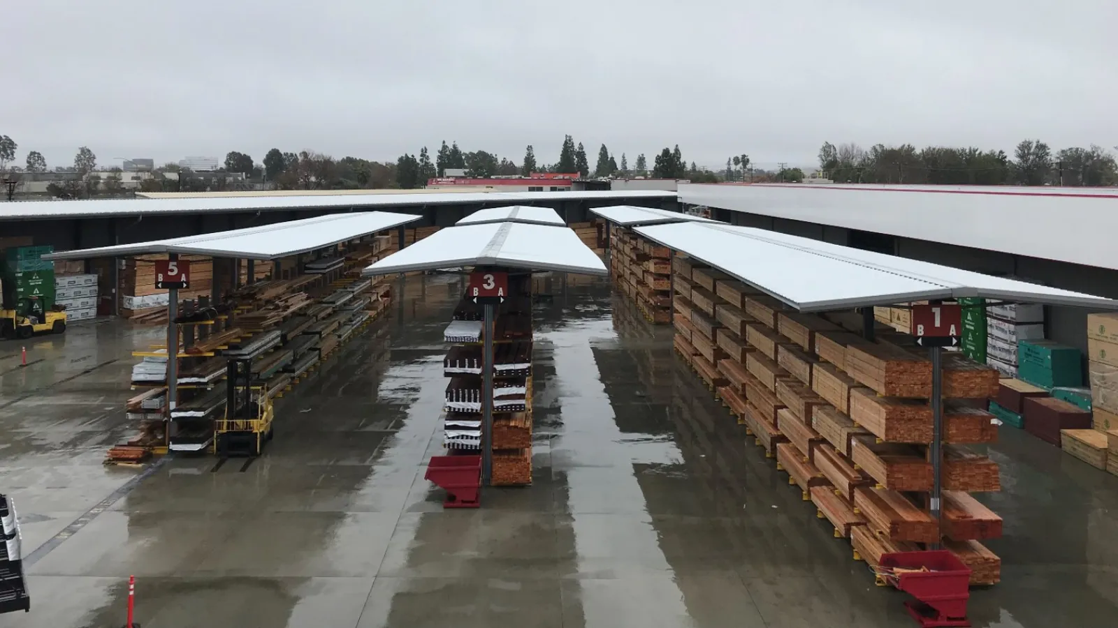 Outdoor lumber yard with stacks of wood and metal roofing under cloudy sky on wet concrete ground