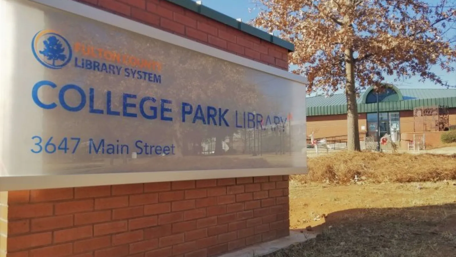 College Park Library sign on brick base with building and autumn tree in background under clear sky