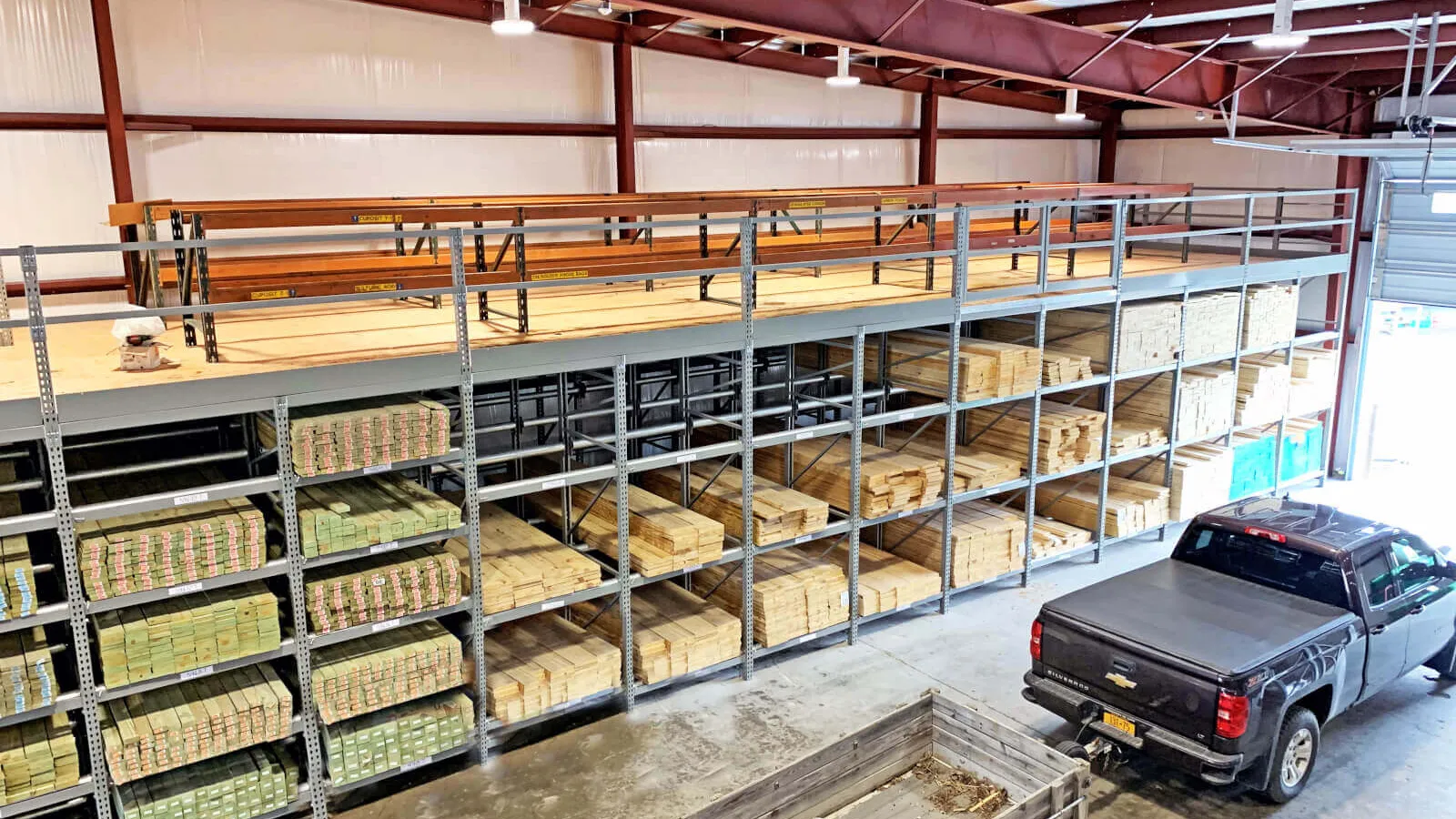 Warehouse interior with metal shelving stocked with lumber and a black pickup truck with trailer inside.