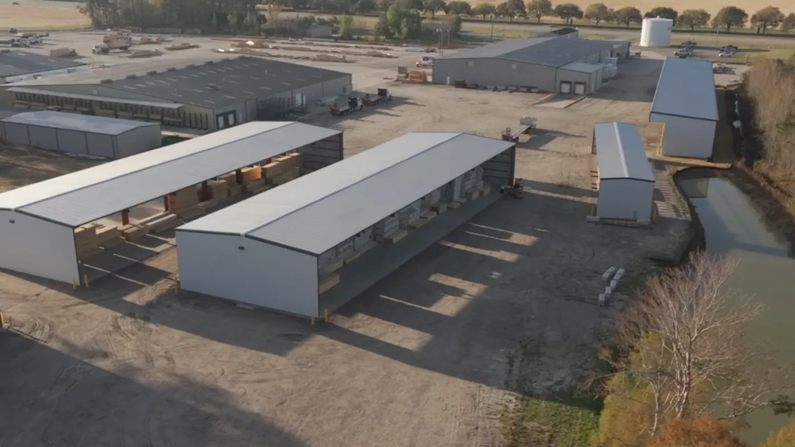 Aerial view of industrial warehouse buildings and storage facilities on a large dirt lot next to a canal.