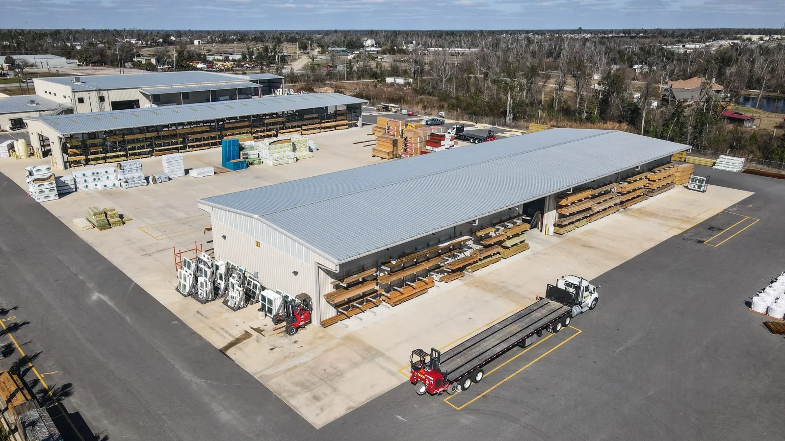 Aerial view of a large lumber storage yard with metal roofing, stacks of wood, and trucks parked on concrete.