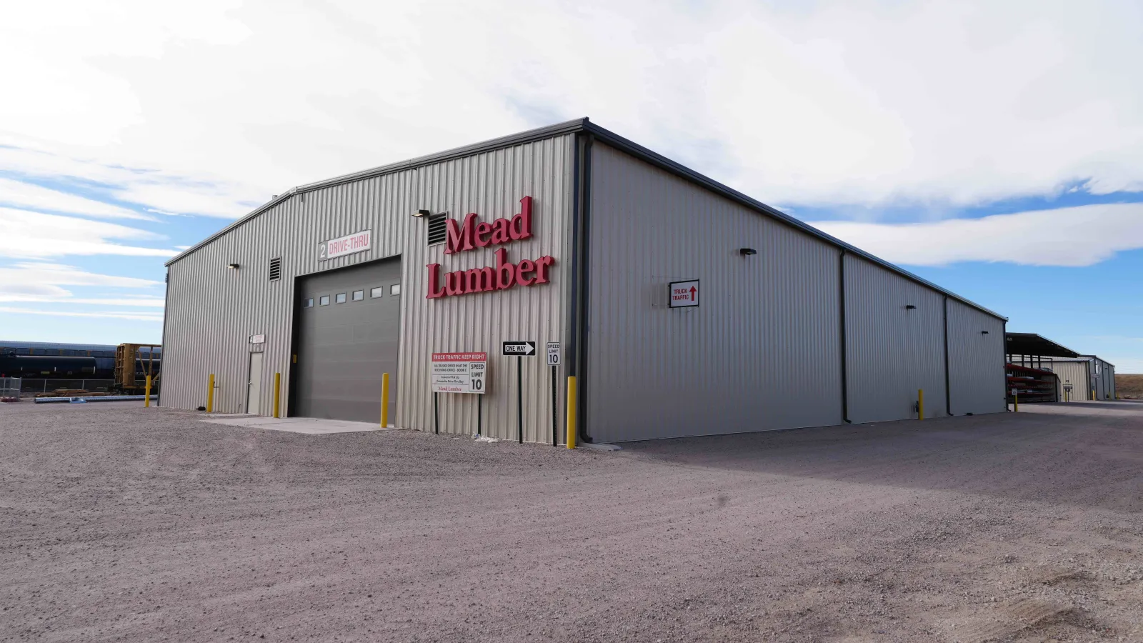 Large gray metal warehouse building with Mead Lumber signage and drive-thru entrance under a cloudy sky