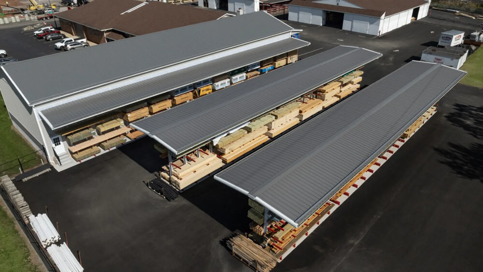 Aerial view of a lumber yard with covered storage racks and multiple buildings on a paved lot under clear sky.