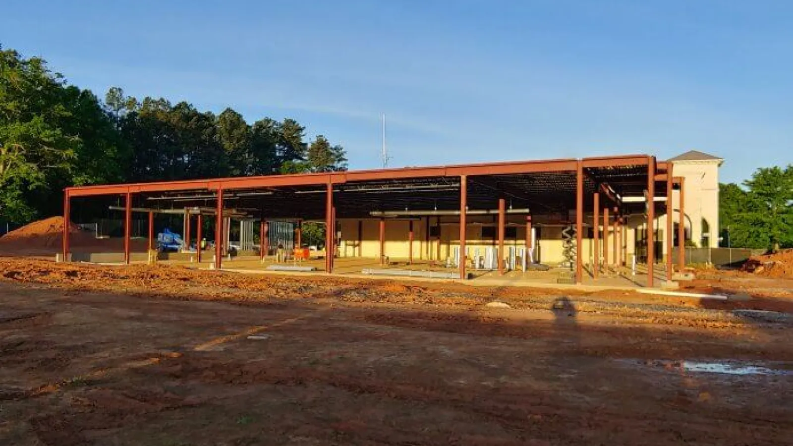 Steel framework of a building under construction surrounded by dirt and trees on a sunny day