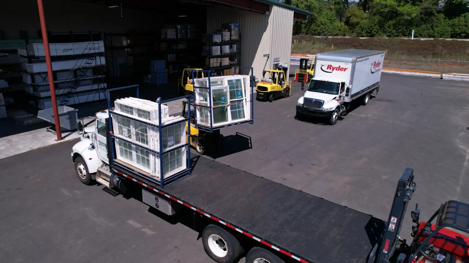 Flatbed truck loaded with window frames, forklifts moving glass pallets near warehouse and delivery truck outside.