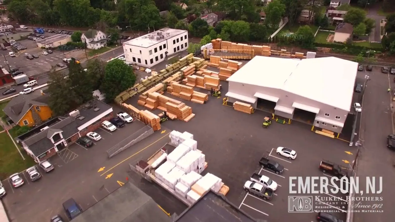 Aerial view of a lumber yard in Emerson, NJ with stacked wood materials, a white warehouse, and parked vehicles.