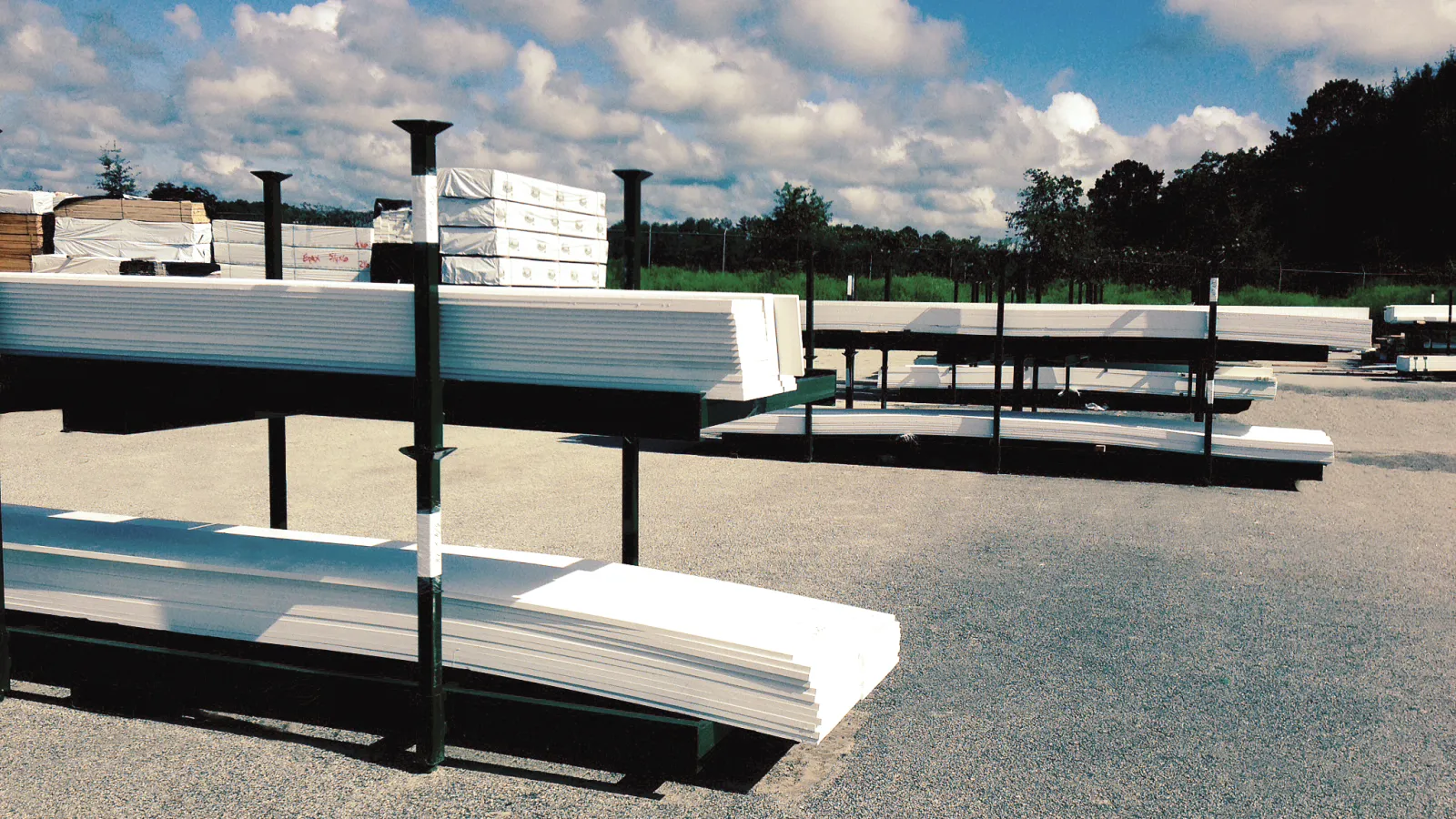 Stacks of white building materials neatly arranged on outdoor metal racks under a bright blue sky with clouds.
