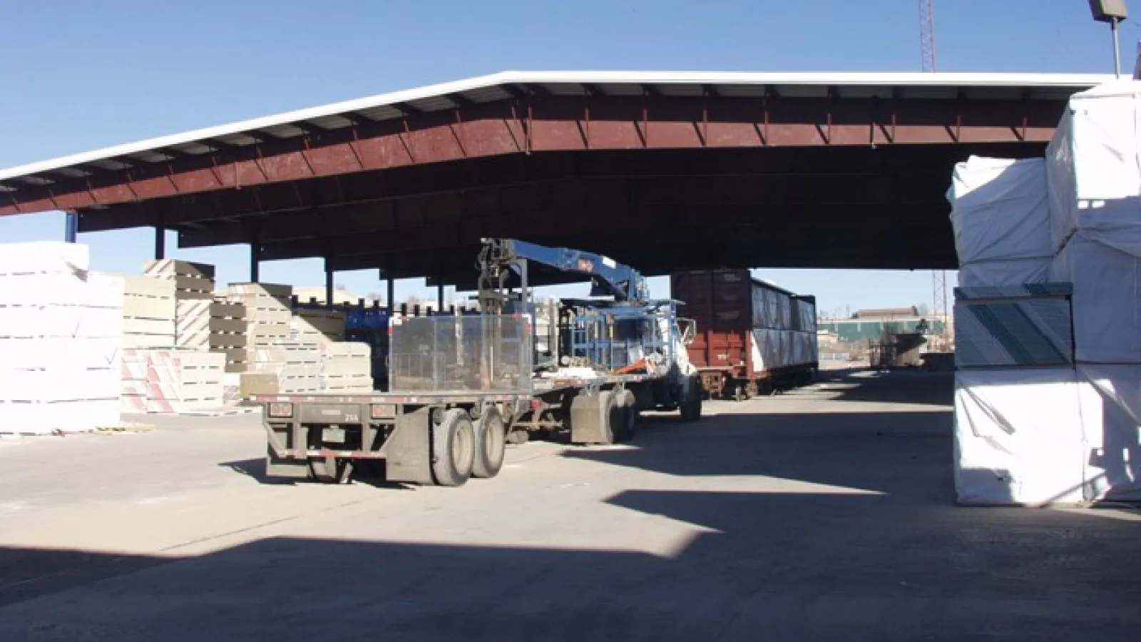 Industrial warehouse with flatbed trucks and stacks of construction materials under a large metal roof