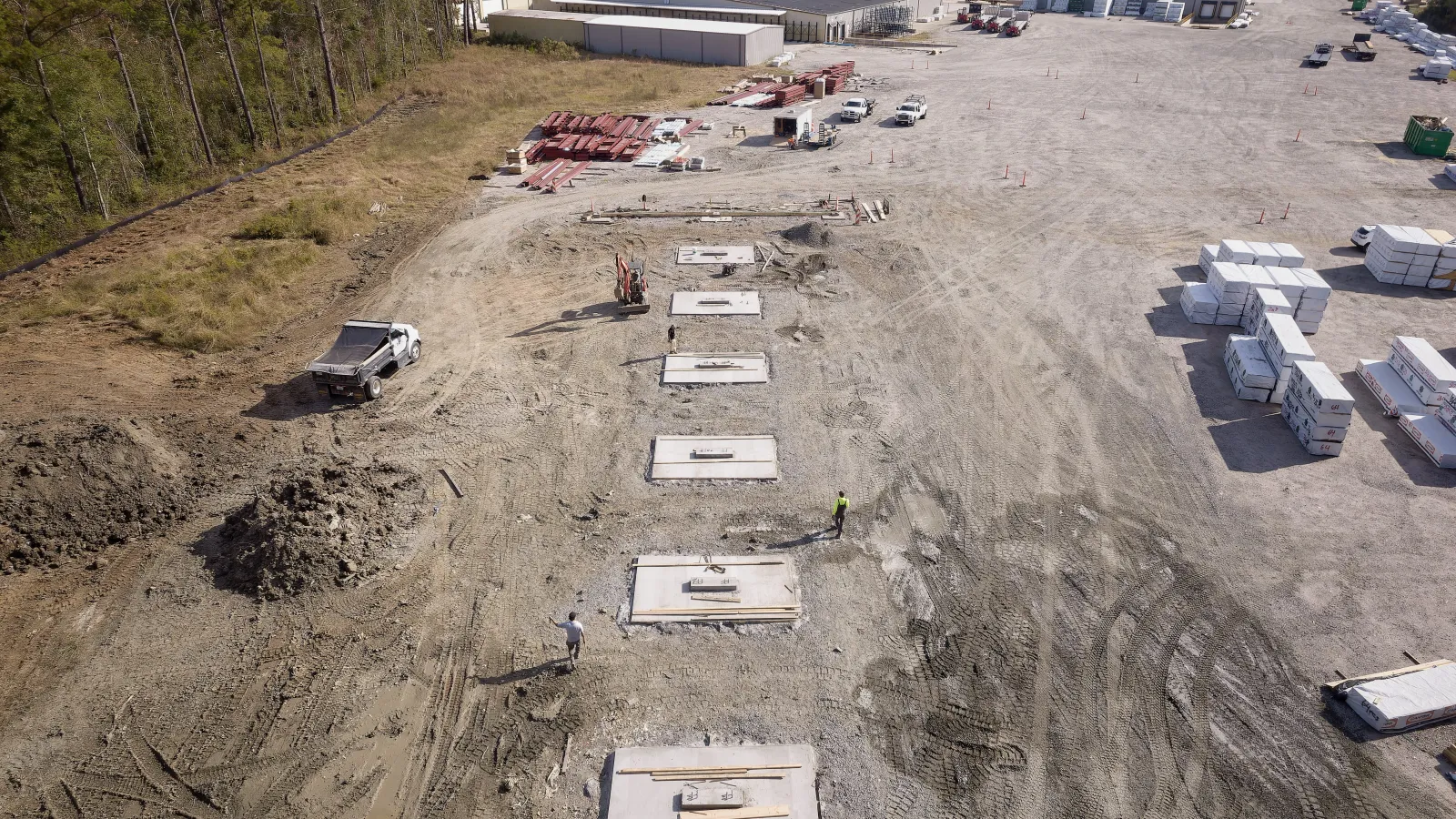 Aerial view of a construction site with concrete slabs, workers, machinery, and building materials on dirt ground.