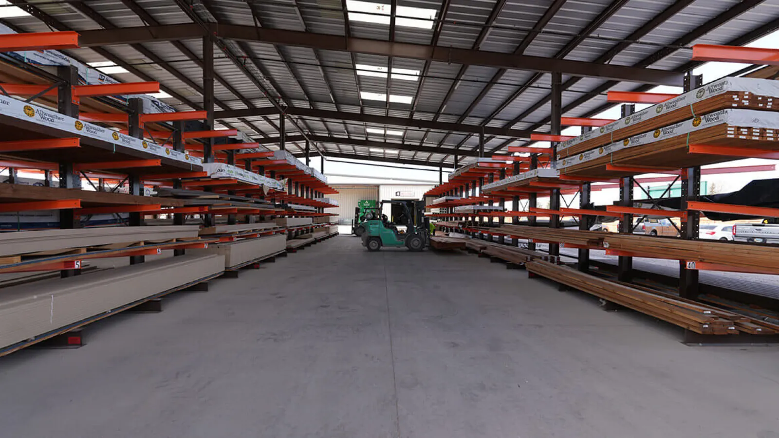 Warehouse interior with metal and wood materials on orange racks and a green forklift in the center aisle.