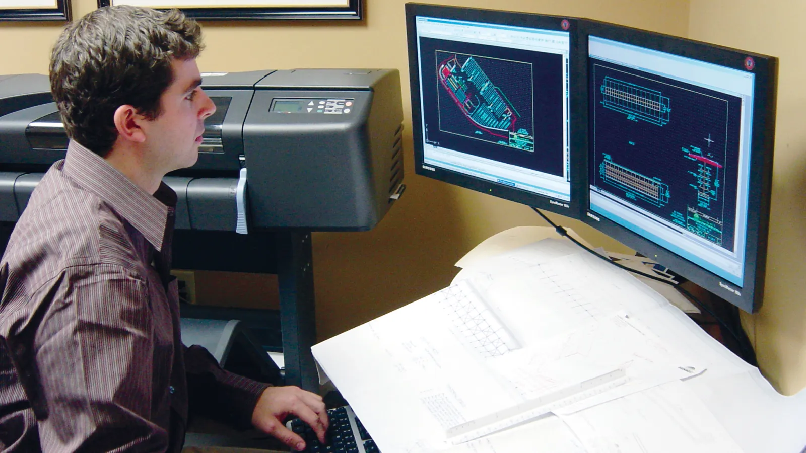 Engineer working on dual monitors with architectural blueprints and CAD designs in office workspace.