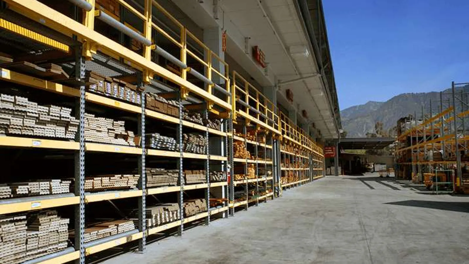 Outdoor warehouse with long metal shelves stocked with various lumber and wood materials under clear blue sky.