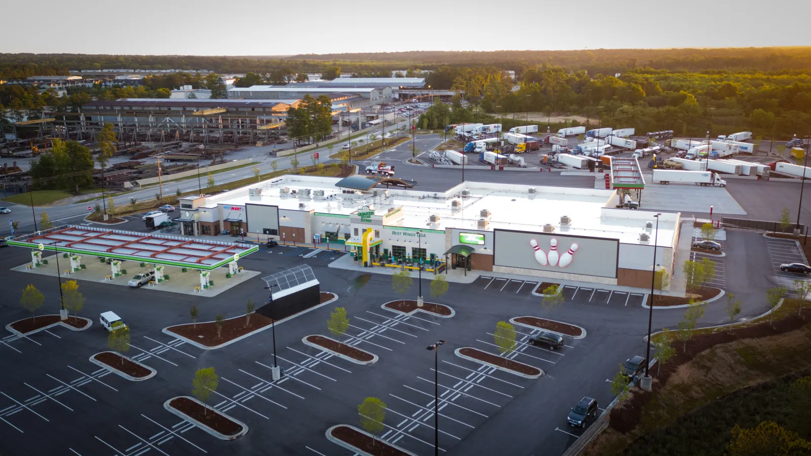 Aerial view of a large bowling alley and gas station with a mostly empty parking lot at sunset.