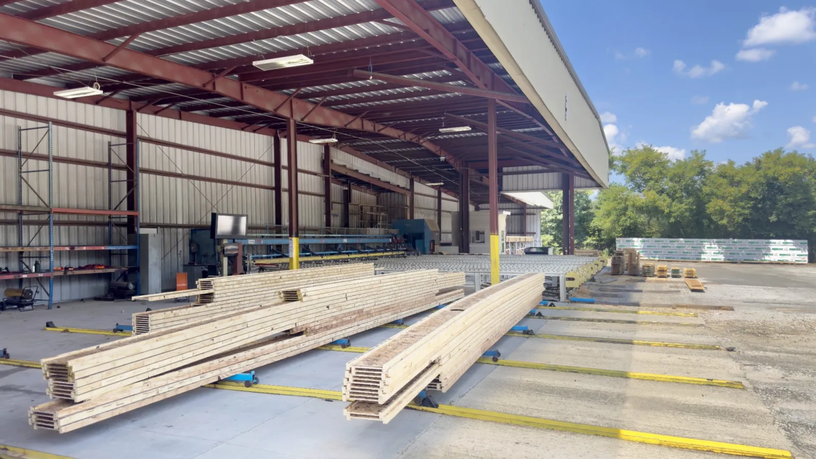 Outdoor lumber storage area with stacked wooden beams under a metal roof structure on a sunny day.