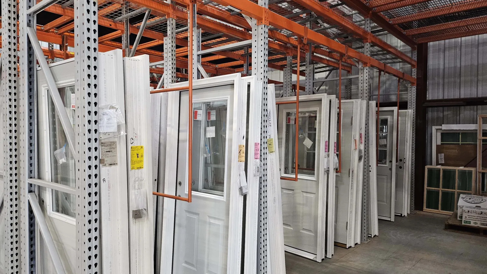 Warehouse interior with metal shelves holding window frames, glass panes, and wooden materials under warm lighting.