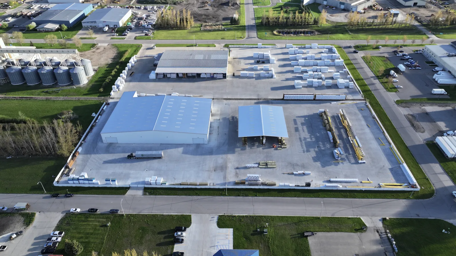 Aerial view of an industrial warehouse with storage areas and trucks in a suburban area under clear skies.