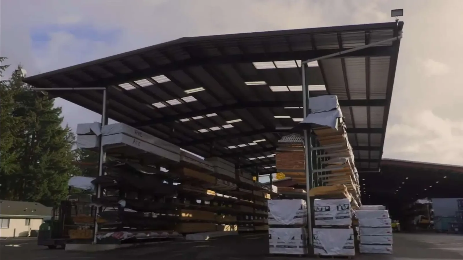 Outdoor lumber storage rack under metal canopy with stacks of wrapped and unwrapped wood materials.