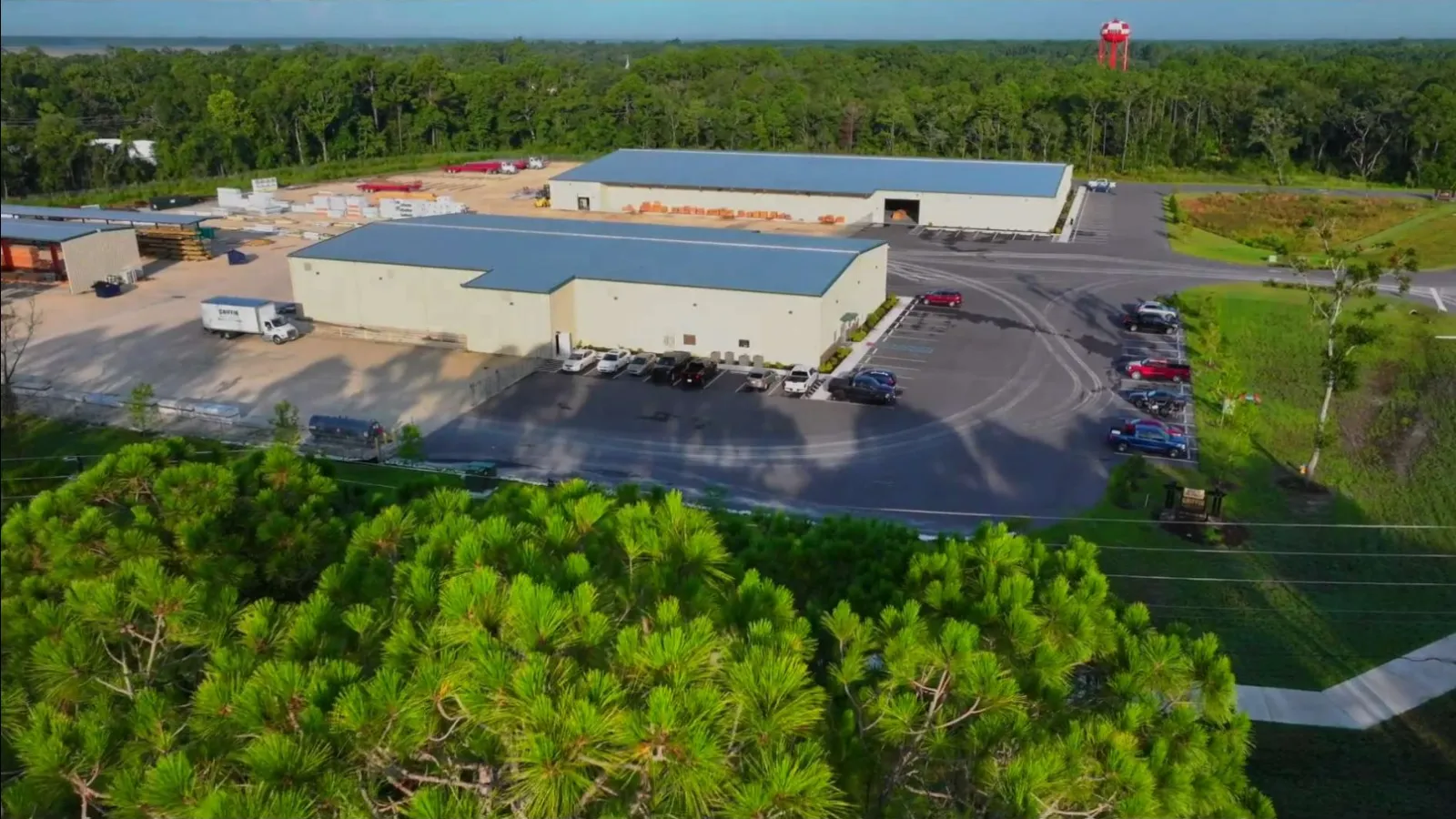 Aerial view of industrial warehouse with parking lots surrounded by greenery under clear sky.