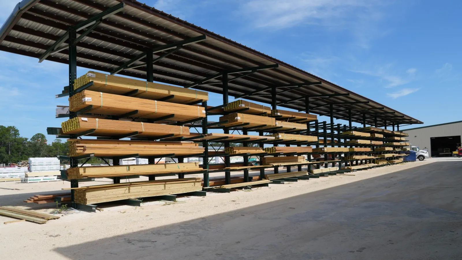Outdoor lumber yard with multiple stacked wood planks stored on metal racks under a metal roof against blue sky.