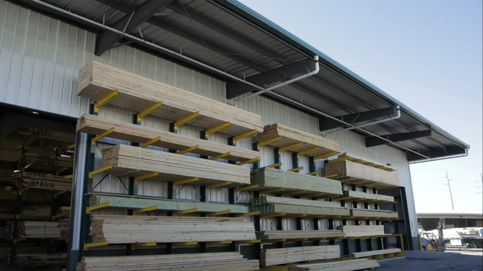 Stacks of lumber neatly organized on racks outside a large warehouse under a clear blue sky.