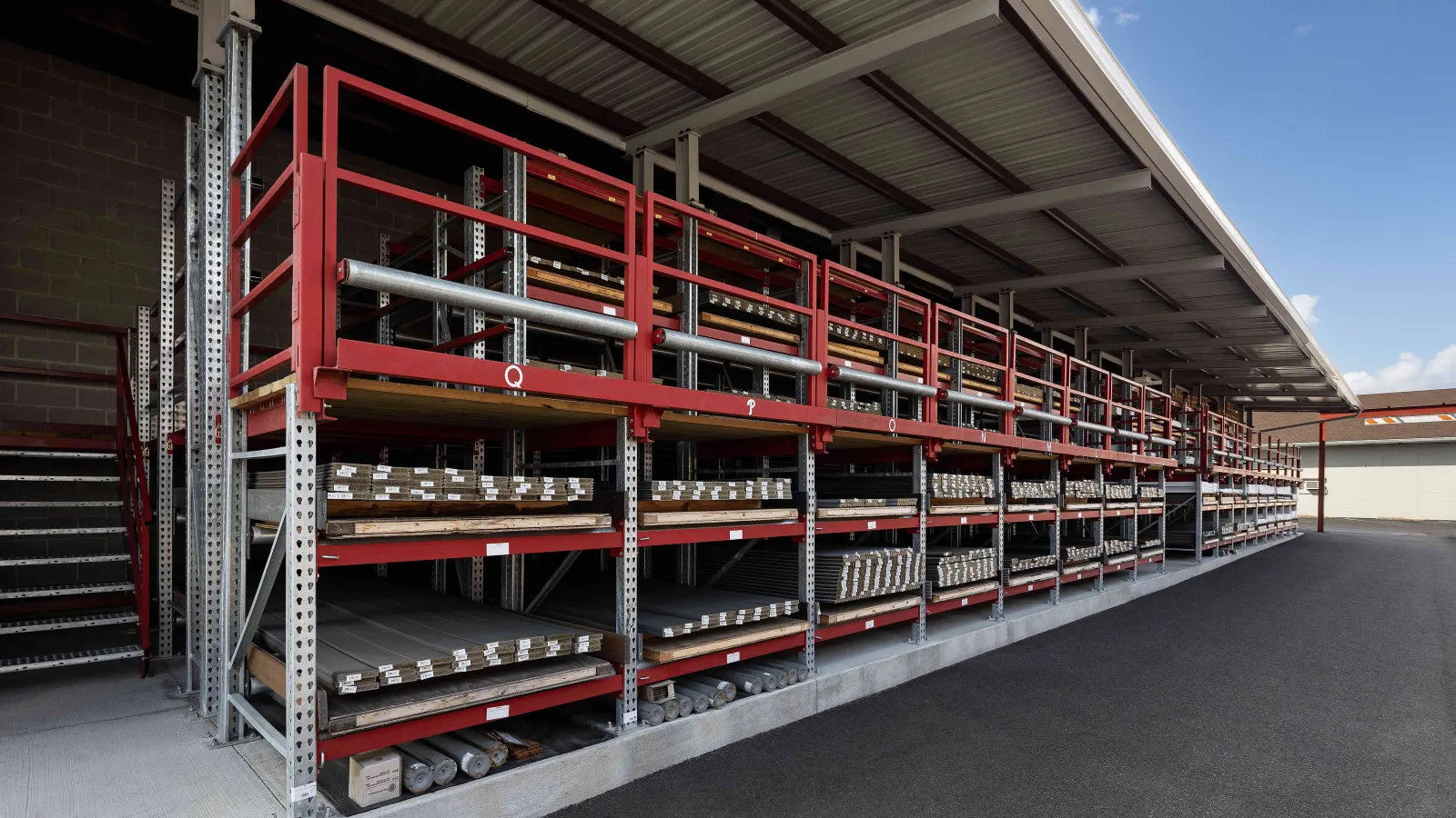 Outdoor industrial storage racks holding metal pipes and materials under a large metal roof structure.