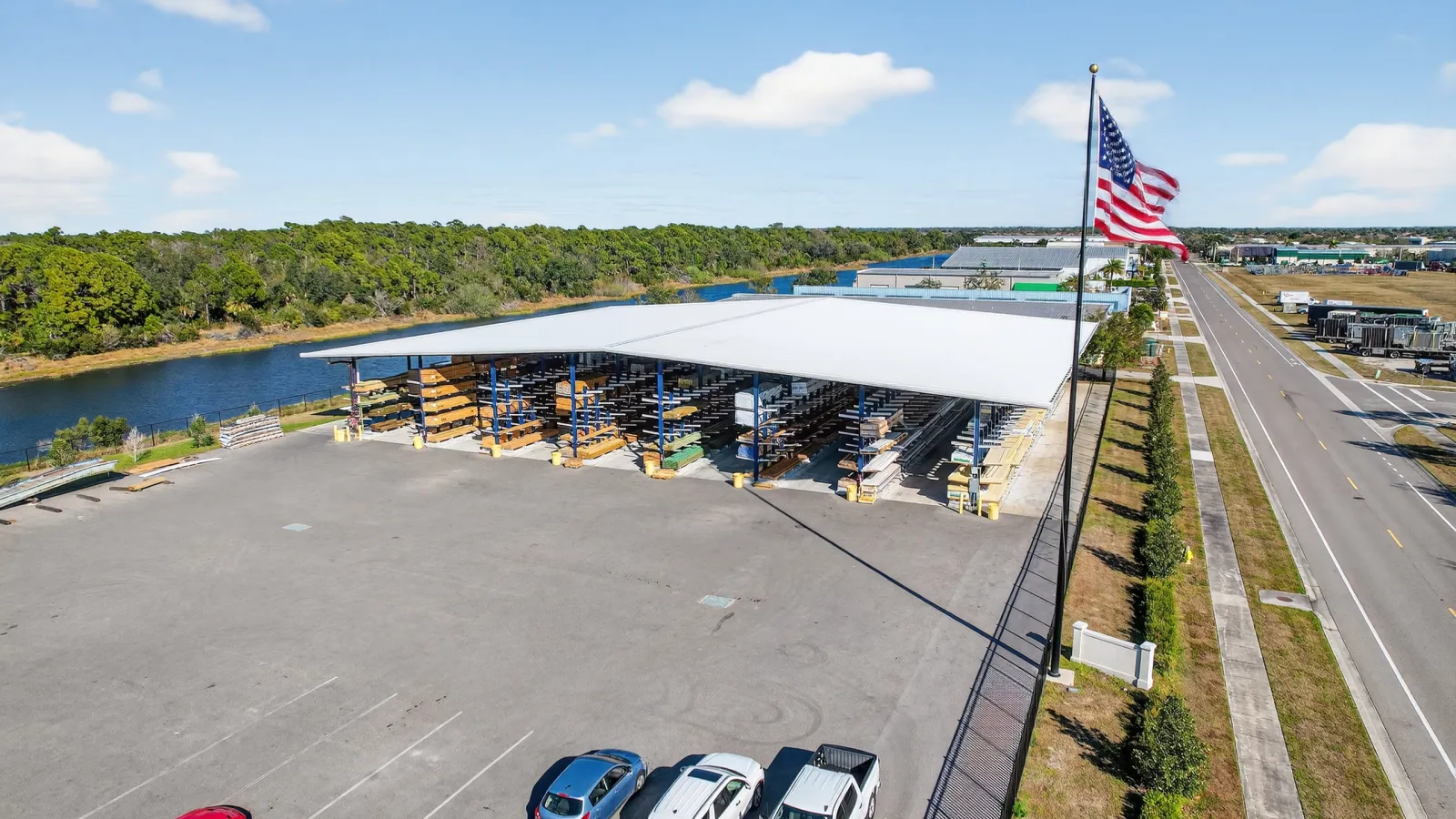 Open-sided warehouse with stored lumber, parked cars, American flag, near road and waterway under clear sky.