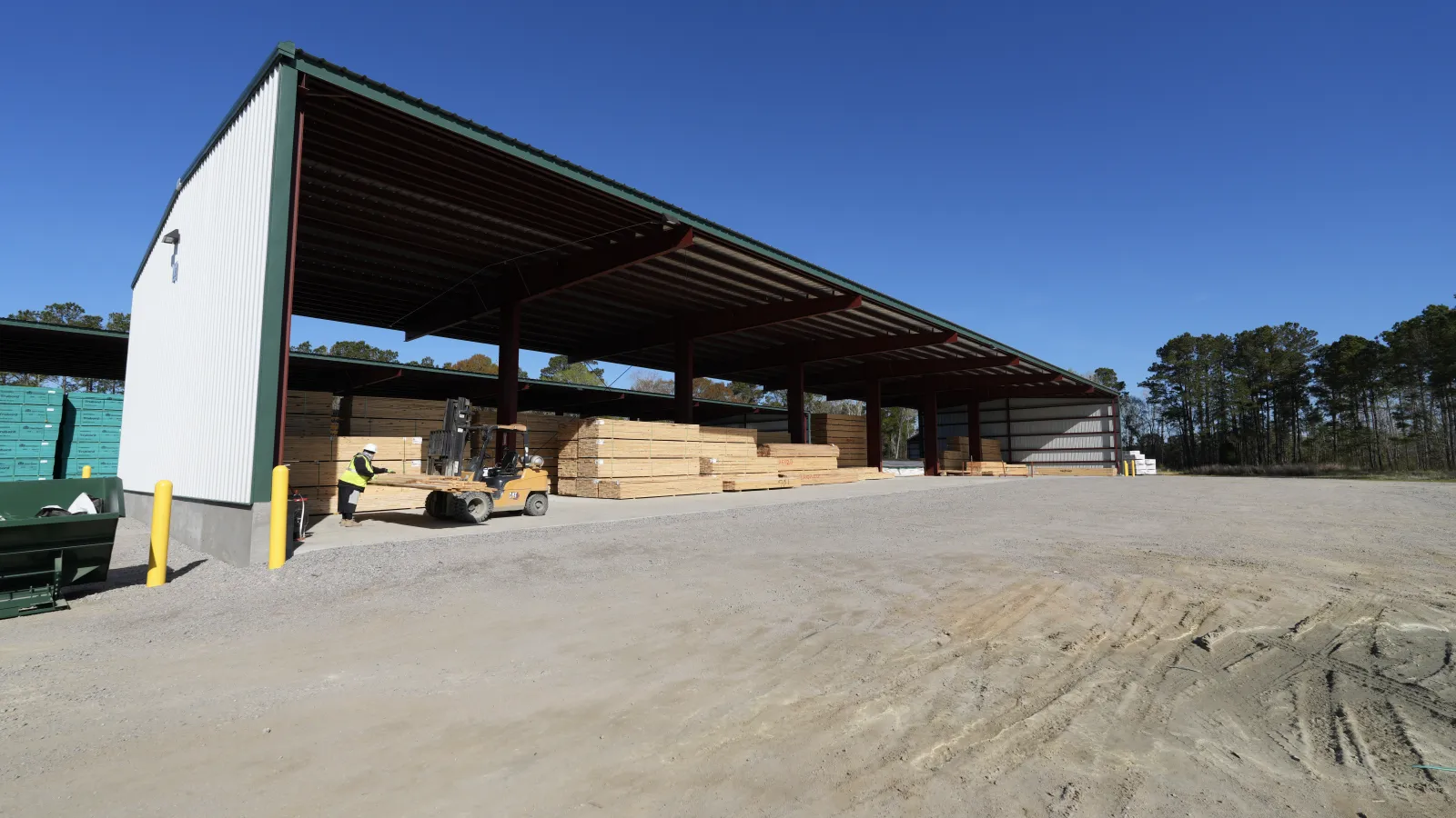 Open lumber yard with stacked wood under large metal roof and forklift operating under clear blue sky.