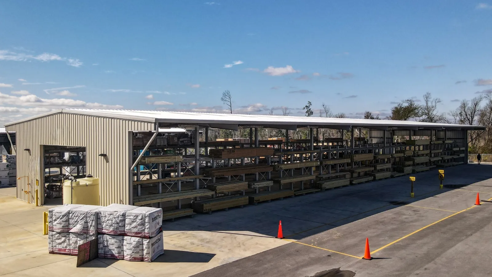 Outdoor lumber storage rack under metal roof next to warehouse with stacked building supplies and cones on concrete.