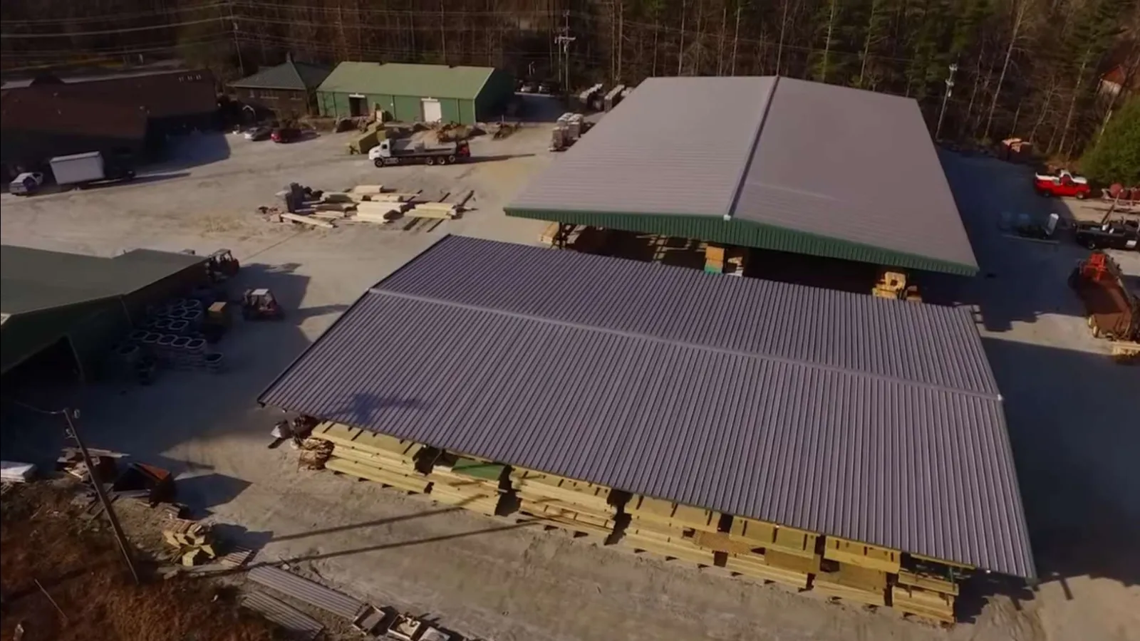 Aerial view of a lumber yard with stacks of wood under metal roofs and surrounding buildings in a forested area.