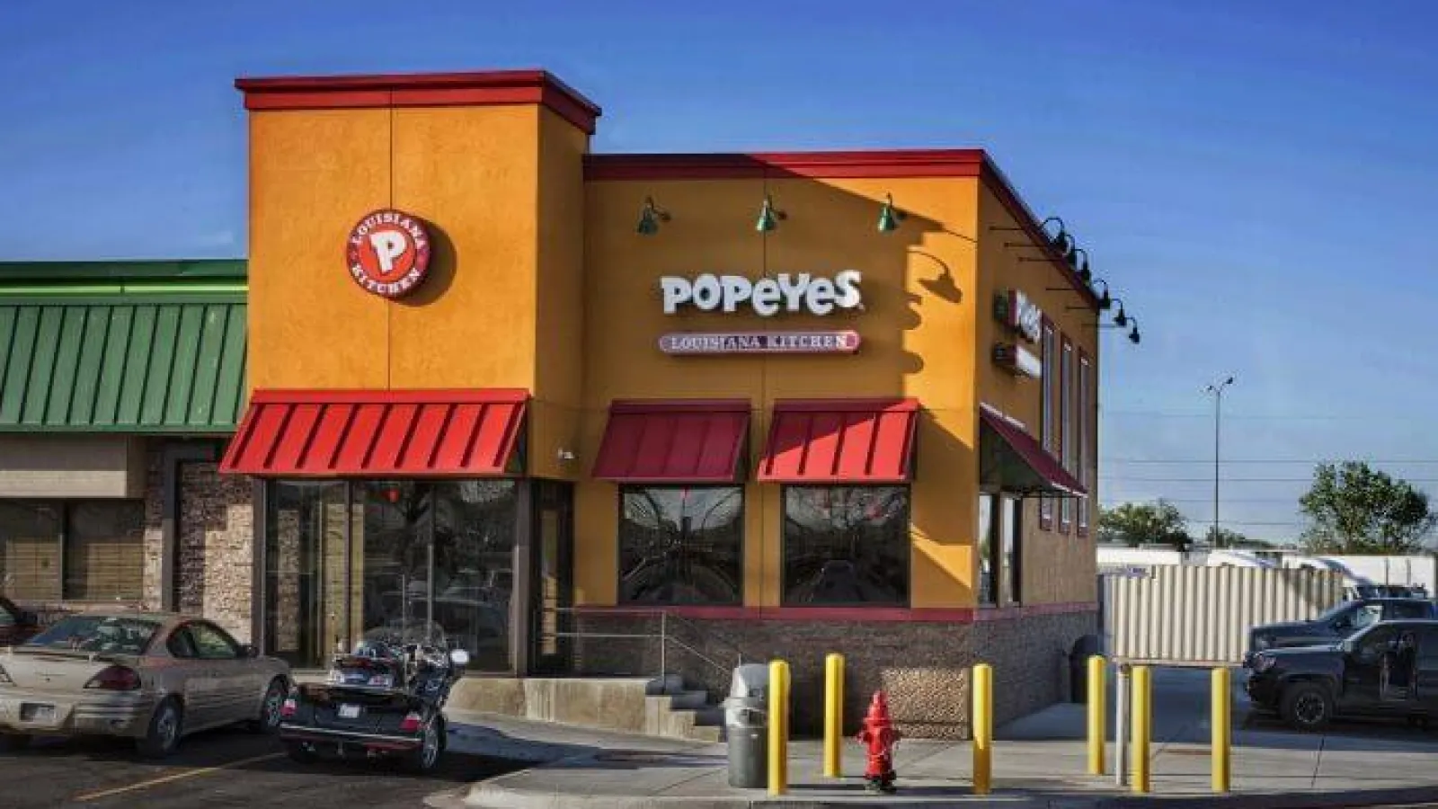 Popeyes Louisiana Kitchen restaurant exterior with orange walls, red awnings, and parked vehicles in front.