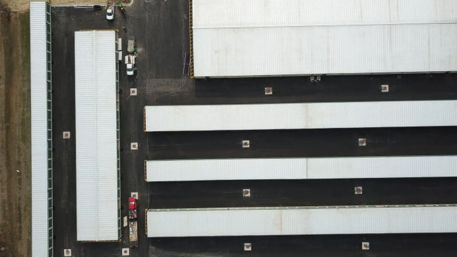 Aerial view of long white-roofed industrial buildings with paved black roads and parking areas around them.