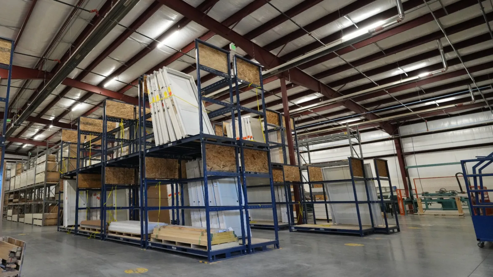Spacious warehouse interior with industrial shelving holding large glass panes and wooden pallets under metal roof beams