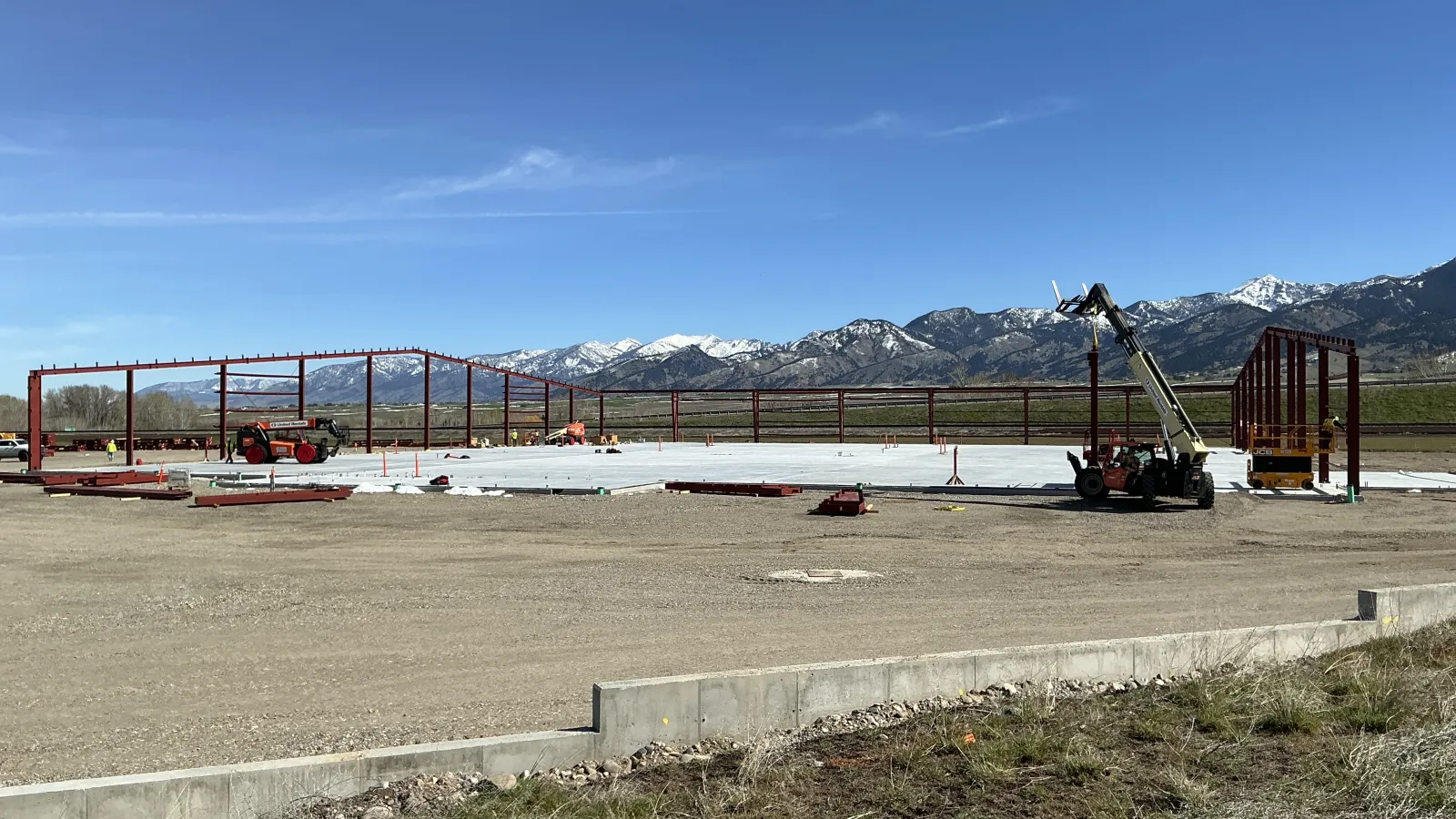 Steel frame structure under construction on concrete slab with mountain range and clear blue sky background.