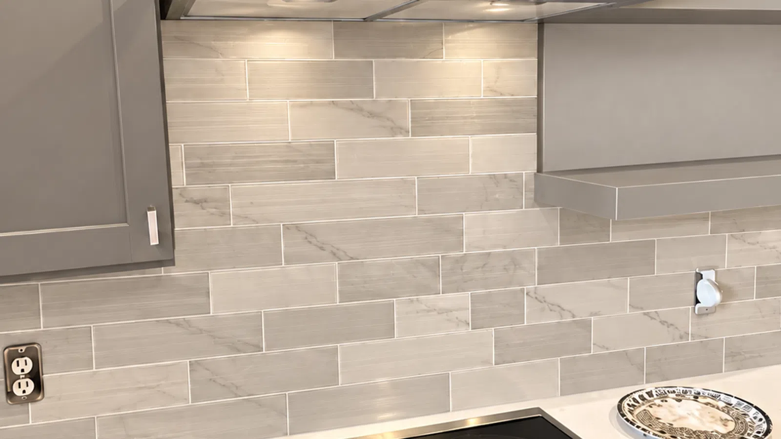 Modern kitchen electric cooktop with stainless steel range hood and gray tiled backsplash in a bright setting