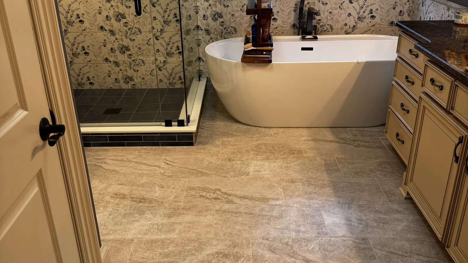 Modern bathroom with freestanding tub, glass shower, floral wall tiles, beige floor tiles, and dark granite countertop.