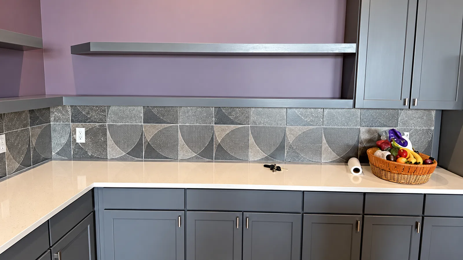 Modern kitchen corner with purple walls, gray cabinetry, geometric tiled backsplash, and a fruit basket on countertop
