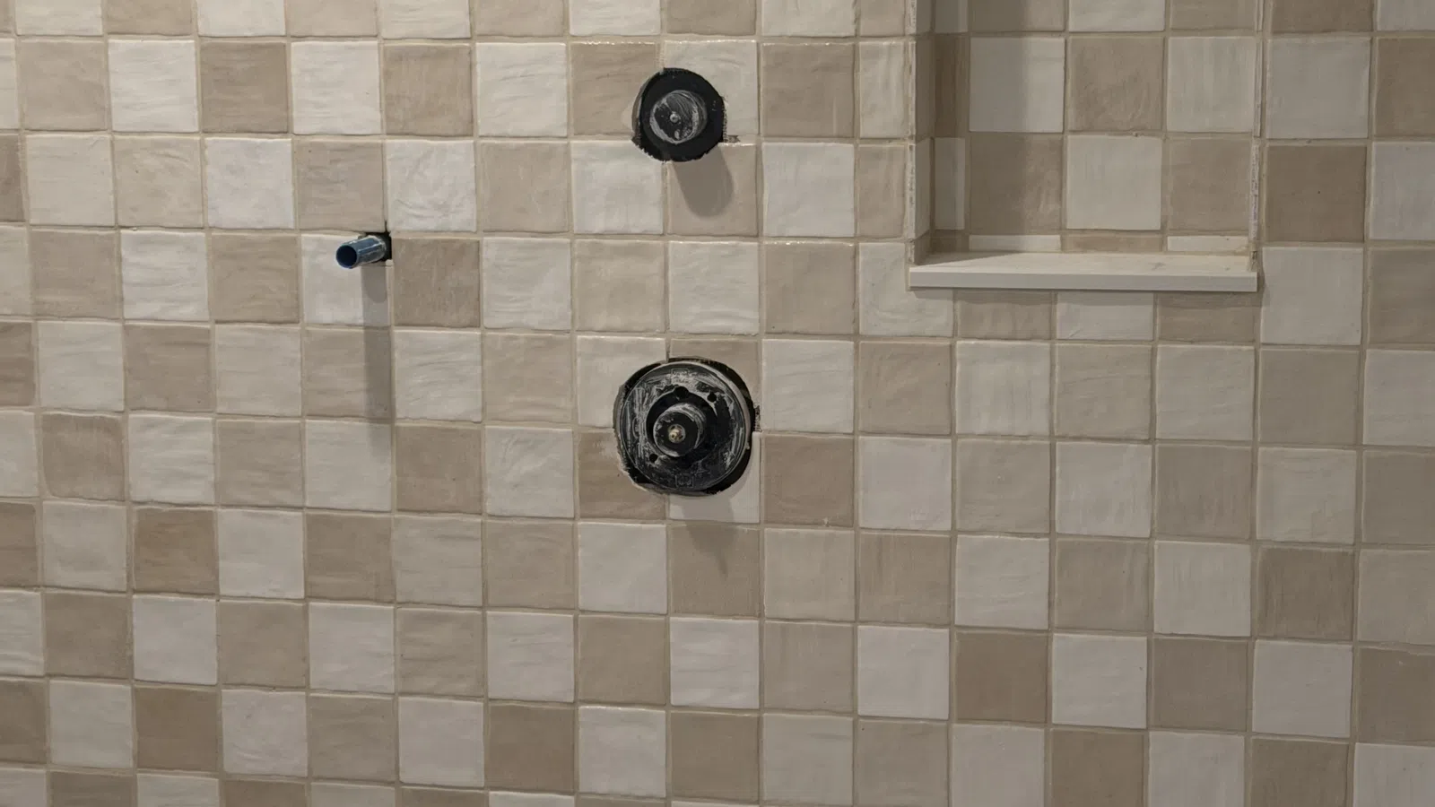 Beige and cream checkered tiled shower wall with built-in niche and exposed plumbing fixtures.