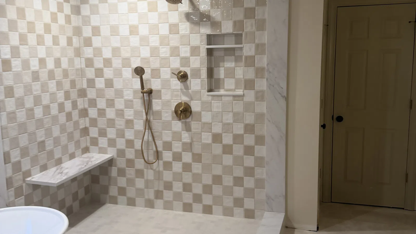 Modern walk-in shower with beige and white checkerboard tiles, bronze fixtures, built-in shelf, and marble bench.