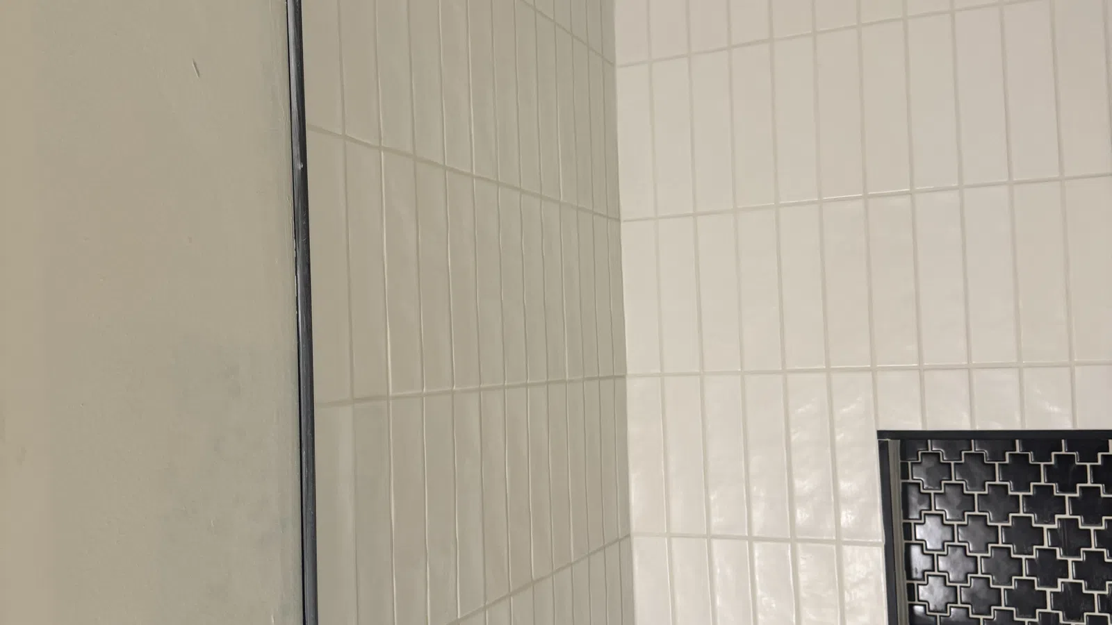 White rectangular wall tiles with a black patterned vent inset in a corner bathroom wall.